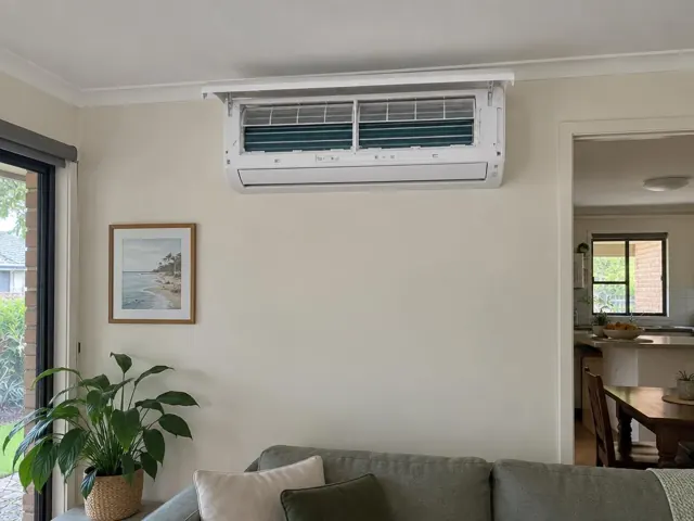 Open split system indoor unit high on a cream wall in a living room, with plants and doorway in Merrimac