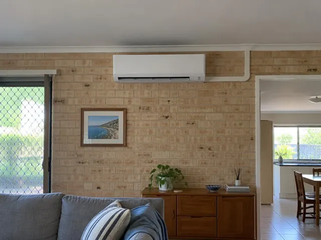 Wall-mounted split system above a brick feature wall in a living room with timber cabinet and doorway in Burleigh Waters